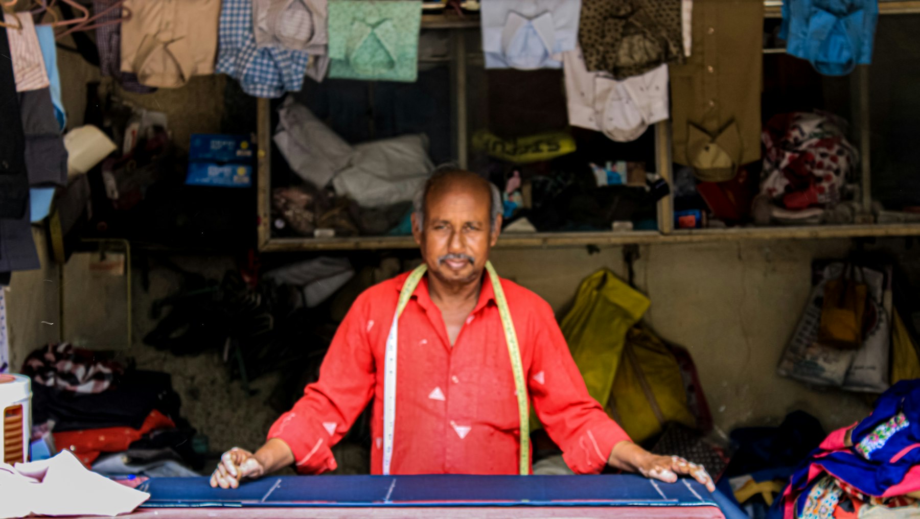 A tailor and shopkeeper in a red shirt with a measuring tape stands behind his counter, shirts stacked on the shelves behind him