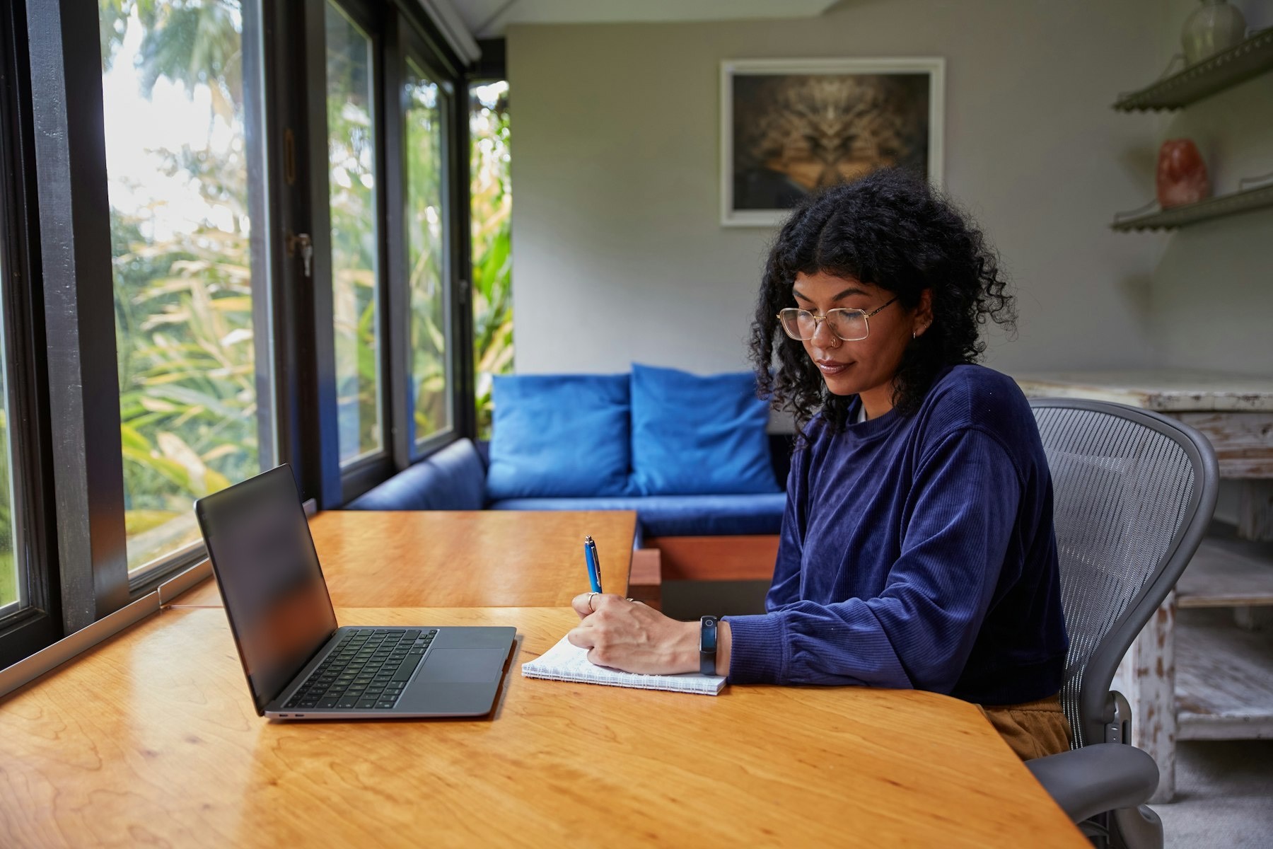 A woman writes in a notebook at a wooden table beside an open laptop, in a bright room with tall windows onto green foliage