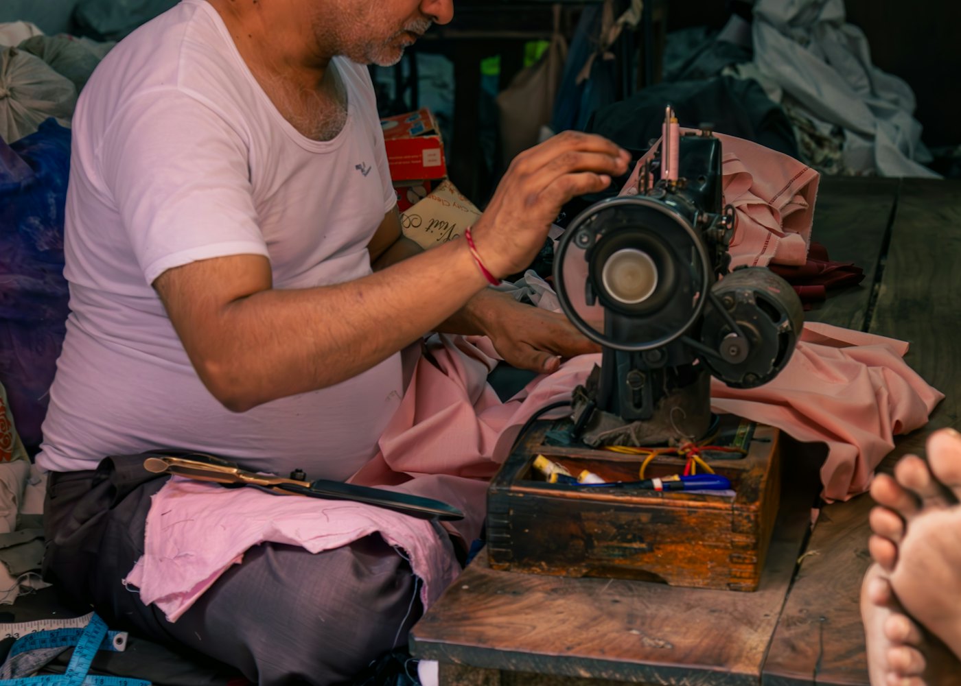 A tailor works at a treadle sewing machine, guiding cloth under the needle